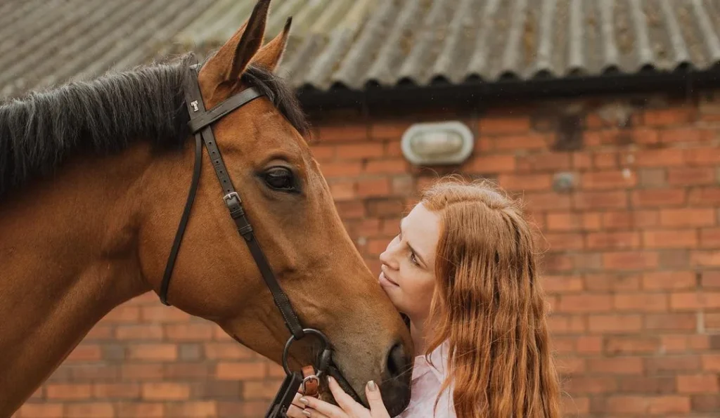 girl looking at her brown horse while posing for horse portrait photography with a brick wall from the stable in the background