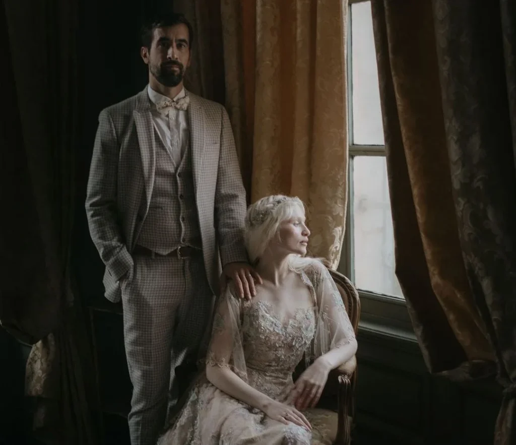 wedding couple standing and sitting to pose for their wedding photography, natural lighting by a window, woman looking outside, man laying his arm on her shoulder
